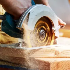 Main d'un artisan guidant une scie circulaire. Le sens de rotation de la lame de scie circulaire projette de la sciure.