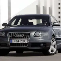 Elegancki, ciemnoszary Audi A6 C6 z przyciemnianymi szybami i chromowanymi felgami, zaparkowany przed nowoczesnym budynkiem.