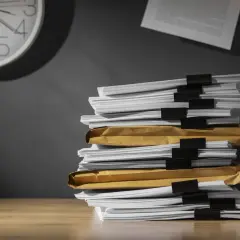 Una pila di documenti su una scrivania, con una nota appiccicata al muro e un orologio che segna il tempo. Chissà ogni quanto si aggiorna il dvr con tutto questo lavoro arretrato.