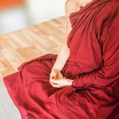 Osoba w czerwonej szacie medytuje, siedząc na poduszce obok drewnianej ławki.