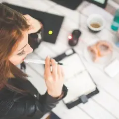 Kobieta pracuje przy laptopie, notuje w zeszycie. Na stole kawa, pączek i kwiaty. Może to praca zdalna, która daje dodatek za prace w weekendy.