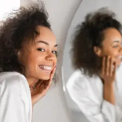 Lächelnde Frau mit lockigem Haar, die sich im Spiegel betrachtet. Ihre Haut strahlt, vielleicht dank Retinol jeden Tag.