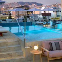 Piscina en la azotea del Barceló Raval con vistas a la ciudad al atardecer. Tumbonas, sofás y luces crean un ambiente acogedor.