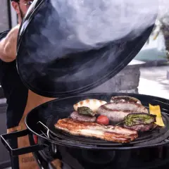 Un homme soulève le couvercle d'un barbecue fumant, révélant une sélection alléchante de viande au barbecue : saucisses, steaks, lard et légumes grillés.