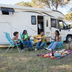 Familie genießt das Zelten mit Kindern. Ein Mann spielt Gitarre, während die anderen singen und lachen. Ein Picknickkorb und Obst liegen auf einer Decke.