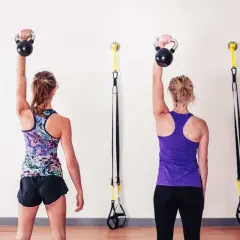 Dwie kobiety ćwiczą z kettlebellami, wykonując rotatory barku. W tle wiszą taśmy TRX.