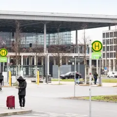 Przystanek autobusowy z zielonym autobusem, ludźmi z bagażami i nowoczesnym budynkiem w tle.