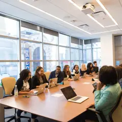 A diverse group of professionals engage in government leadership training, collaborating around a conference table with laptops and tablets.
