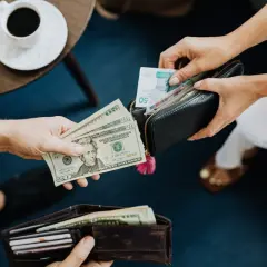Dwie osoby wymieniają się pieniędzmi. Jedna trzyma dolary, druga wkłada do portfela banknoty. Co lepiej zabrać do Turcji euro czy dolary?