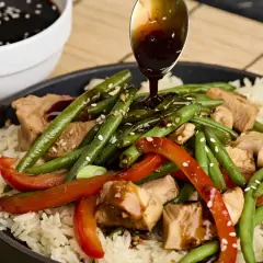 A spoon drizzles teriyaki sauce over a dish of rice, chicken, green beans, and red peppers, garnished with sesame seeds.