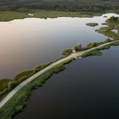 Droga przecinająca jezioro, otoczona zielenią i lasem. W oddali mały domek, jakby cyców na wodzie.