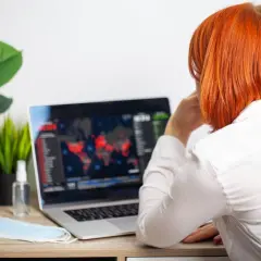 Kobieta z rudymi włosami patrzy na ekran laptopa z mapą świata i danymi. Obok leży maseczka i płyn do dezynfekcji.