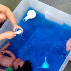 Niños de 3 a 4 años disfrutan de juegos acuáticos en el colegio, explorando con una muñeca y recipientes.