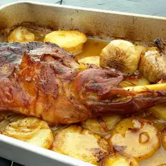 Paletilla de cordero al horno dorada y jugosa, acompañada de patatas asadas y ajos tiernos en una bandeja.