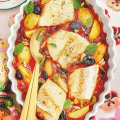 Piatto di baccalà alla romana in teglia con pomodoro, patate, olive e pinoli, guarnito con basilico fresco.