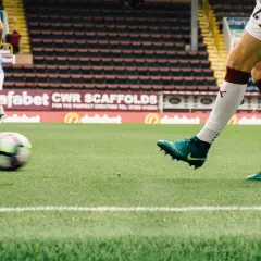 Piłkarz w białych spodenkach i zielonych butach kopie piłkę na boisku. W tle trybuny stadionu, charakterystyczne dla angielskich klubów piłkarskich.