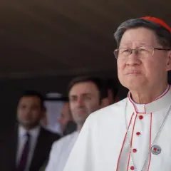 Kardinal Tagle, mit Brille und roter Pileolus, trägt ein weißes Gewand mit roten Akzenten. Im Hintergrund unscharfe Männer.