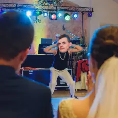 Zabawny moment podczas oczepin weselnych. Pan młody tańczy z wielką głową na szyi, a para młoda obserwuje go z uśmiechem.