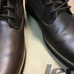 Brązowe, skórzane buty z ciemnymi sznurówkami. Skóra na jednym bucie jest lekko pognieciona, co może sugerować potrzebę dowiedzenia się, jak wyprostować skórę na butach.