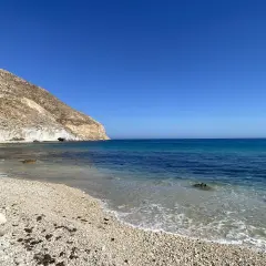 La **playa Peñón Blanco** se extiende bajo un cielo azul intenso. Las olas acarician la orilla de guijarros, mientras una imponente montaña rocosa se alza a la izquierda, con una cueva marina visible en su base.