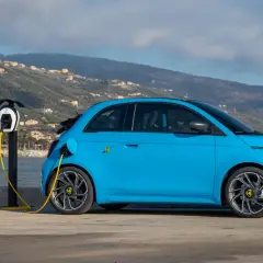 Abarth EV azul cargando junto al mar, con un velero y colinas al fondo.