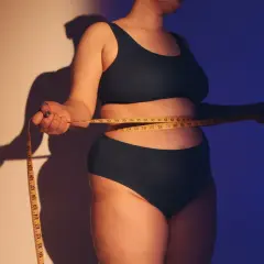 Osoba mierzy talię centymetrem. Ciemne światło podkreśla kształty, wywołując dyskomfort związany z body shaming.