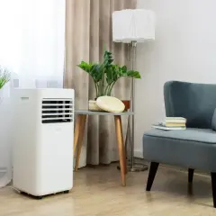 Biały klimatyzator przenośny stoi obok rośliny w wiklinowym koszu. Wygodny fotel z książkami i poduszką dopełnia wnętrze.