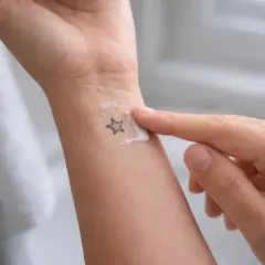 Un doigt applique une crème sur un petit tatouage en étoile sur le poignet. Le tatouage piqué trop profond semble légèrement irrité sous la crème.