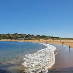 La **playa de Cuchía** se extiende bajo un cielo azul, con gente paseando por la arena dorada y el mar acariciando la costa.