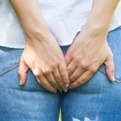Femme tenant ses mains sur son périnée gonflé et douloureux, en jean et chemise blanche.