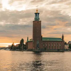 Stockholm Stadshus vid solnedgången, en vy som kunde vara från stockholm 1900. Vatten, båtar och en bro i bakgrunden.