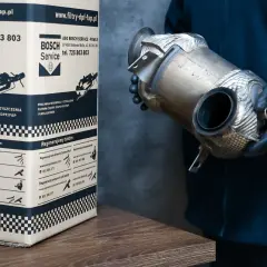 Osoba w rękawiczkach trzyma element układu wydechowego, prawdopodobnie filtr DPF. Obok stoi karton z logo Bosch Service, informujący o regeneracji filtrów DPF w dieslu.