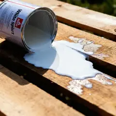Une boîte de white spirit renversée sur des planches de bois. Le liquide blanc s'étale, créant des formes abstraites sur le grain du bois.