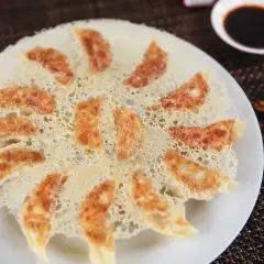 Chrupiące pierożki gyoza ułożone na talerzu, z delikatną, złotą panierką. Obok miseczka sosu sojowego i pałeczki.