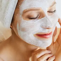 Frau mit weißer Maske im Gesicht, die ihre Haut pflegt. Ein Handtuch ist um ihren Kopf gewickelt.