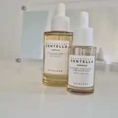 Dwie buteleczki z pipetką zawierające **centella ampoule** od SKIN1004, jedna większa, druga mniejsza, stoją na białej powierzchni.