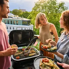 Vänner njuter av en grillfest på balkongen med en bärbar kolgrill. Saftiga biffar och grönsaker grillas medan solen går ner.