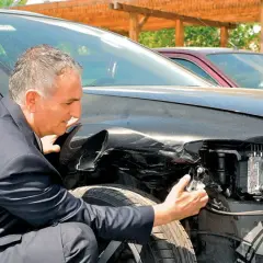 Mężczyzna w garniturze ogląda uszkodzony przód czarnego samochodu. Zastanawia się, **ac co to** za szkody.