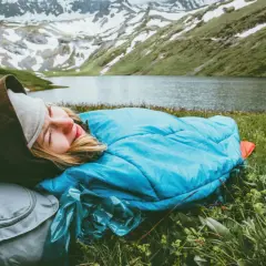 Persona durmiendo en saco azul, con mochila gris, en un prado verde junto a un lago y montañas nevadas. Vivac acampada perfecta.