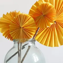 Flores amarillas de papel plegado, como abanicos, en un jarrón de cristal. Parecen flores con manos que se abren.