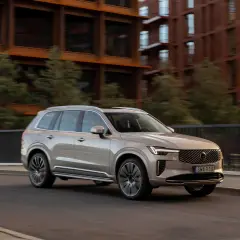 En ljusbeige Volvo SUV kör på en stadsgata. Bakgrunden visar moderna tegelbyggnader och gröna träd. Bilen visar upp olika volvo utrustningsnivåer.
