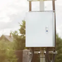 Caja blanca con cierre azul, montada en poste. Su diseño IP44 es para exterior, protegiendo equipos en jardines o patios.