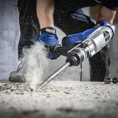 Mocny udar w wiertarce kruszy beton, wzbijając chmurę pyłu. Pracownik w niebieskich rękawicach kontroluje narzędzie.