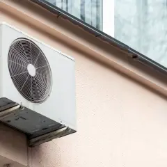 Klimatyzacja w bloku, biała jednostka zewnętrzna zamontowana na jasnoróżowej ścianie, z widocznym wentylatorem i kratką.