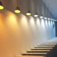 Escalera con luces colgantes que crean un degradado de luz y color, desde cálido hasta frío, iluminando la pared texturizada.