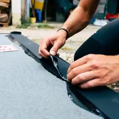Mains habiles en train d'enlever la colle néoprène d'une bande noire sur une surface grise.