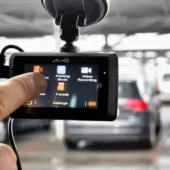 Palec dotyka ekranu wideorejestratora, wybierając opcję "Parking Mode". W tle widać zaparkowane samochody.