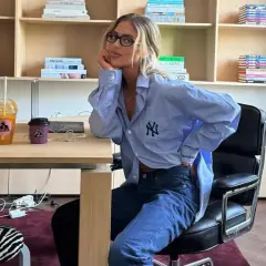 Chica con gafas sentada en una silla de oficina, luciendo un outfit oficina verano hombre: camisa azul oversize y vaqueros.