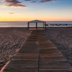 Pasarela de madera hacia una pérgola en la playa de Puerto Rey al atardecer. Sombrillas alineadas al fondo.