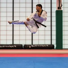 Man i vit dräkt med svart bälte utför ett högt spark i luften under en tae kwon do-träning.
